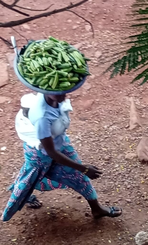 Femme africaine marchant sur un sol en terre, portant un grand plateau rempli de gombos sur la tête, un bébé attaché dans le dos.